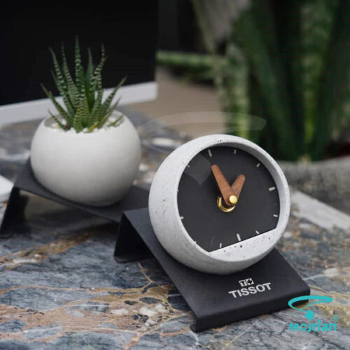 ساعت گوی بتنی تبلیغاتی M8102 با طراحی مینیمال و صفحه سیاه مات. این ساعت روی یک استند فلزی مشکی با حکاکی لوگوی TISSOT قرار گرفته